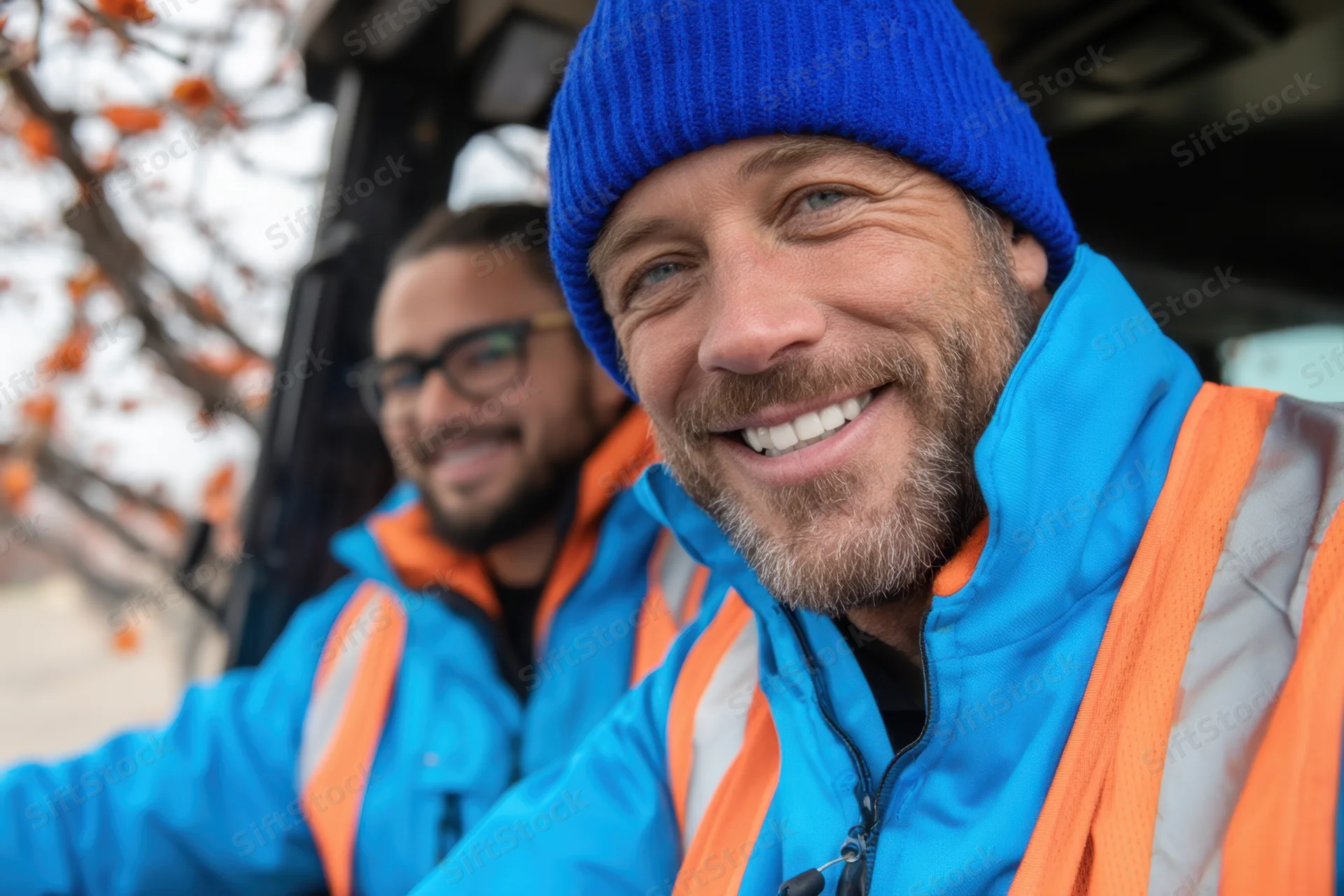 Two men in blue and orange high-visibility jackets smiling while working outdoors