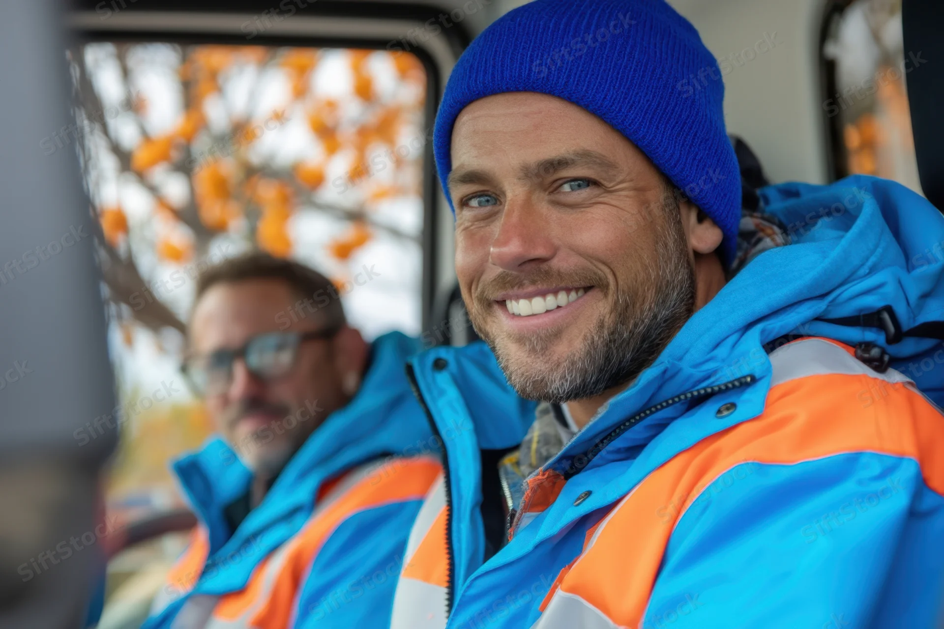 Two men in blue and orange high-visibility jackets smiling inside a vehicle cabin with autumn foliage visible through the window