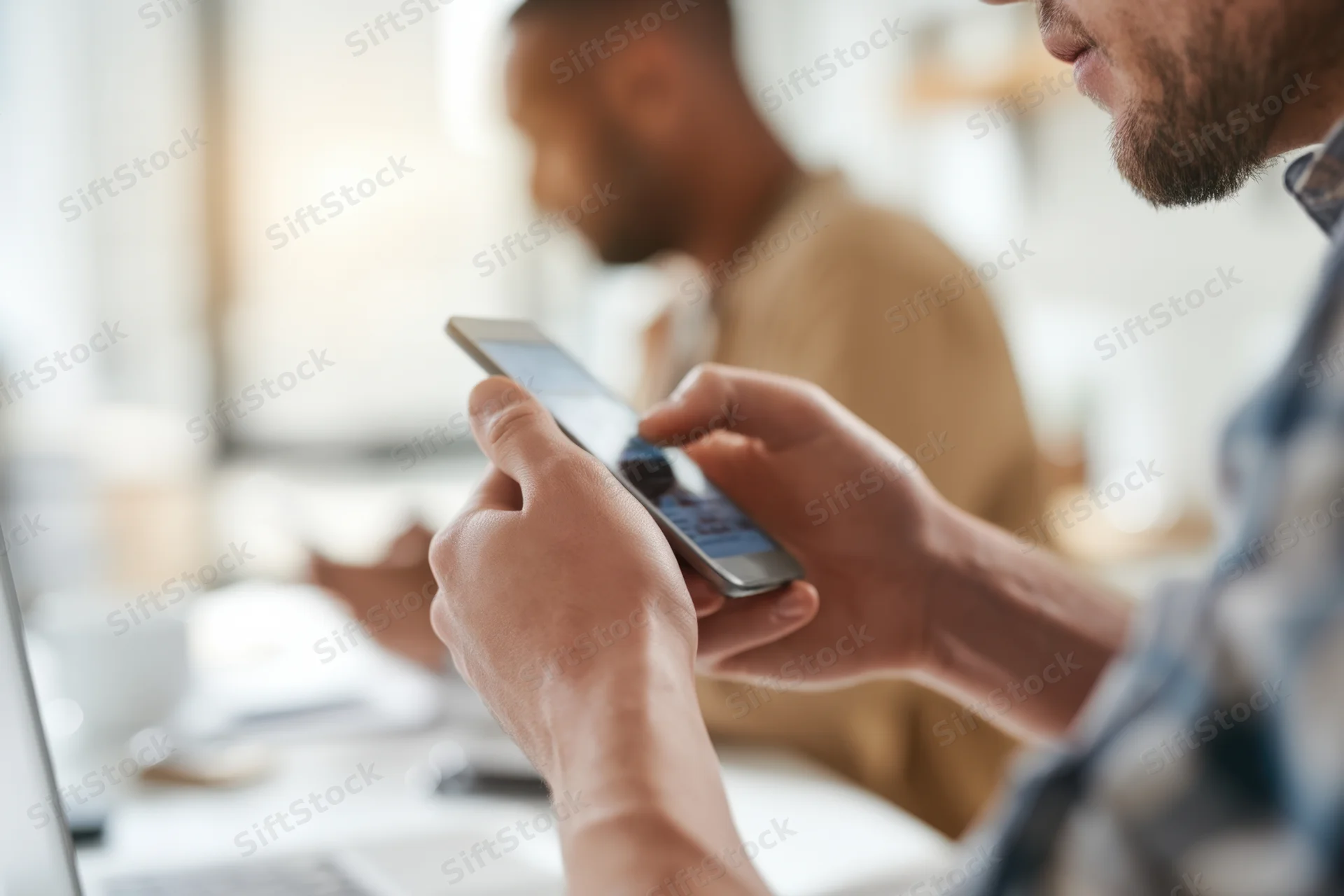 Man using smartphone at desk in modern office with coworker in background