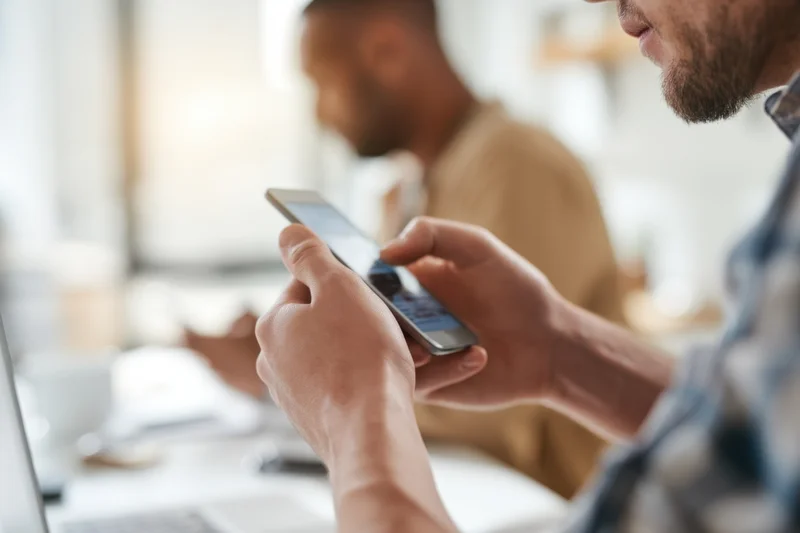 Man using smartphone at desk in modern office with coworker in background