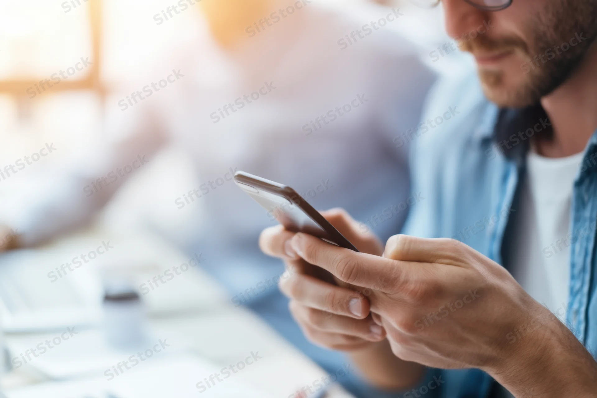 Man typing on smartphone in bright modern office workspace with colleague in background