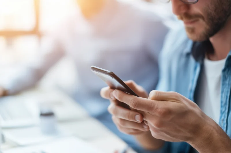 Man typing on smartphone in bright modern office workspace with colleague in background