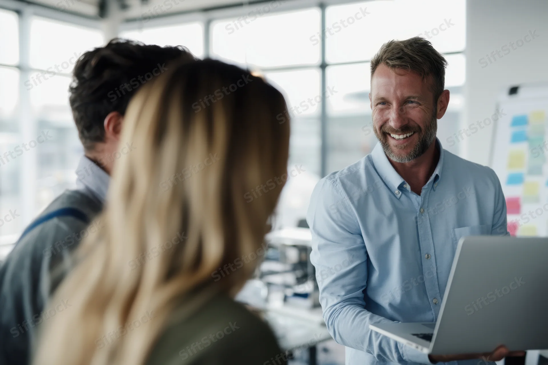Man smiling while holding laptop during team discussion in bright office with large windows