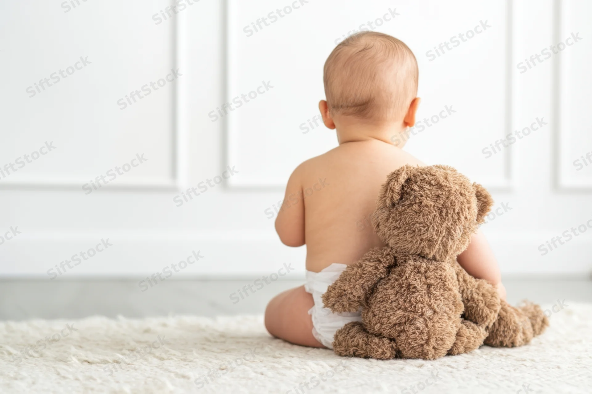 Baby in diaper sitting on light rug cuddling brown teddy bear from back view