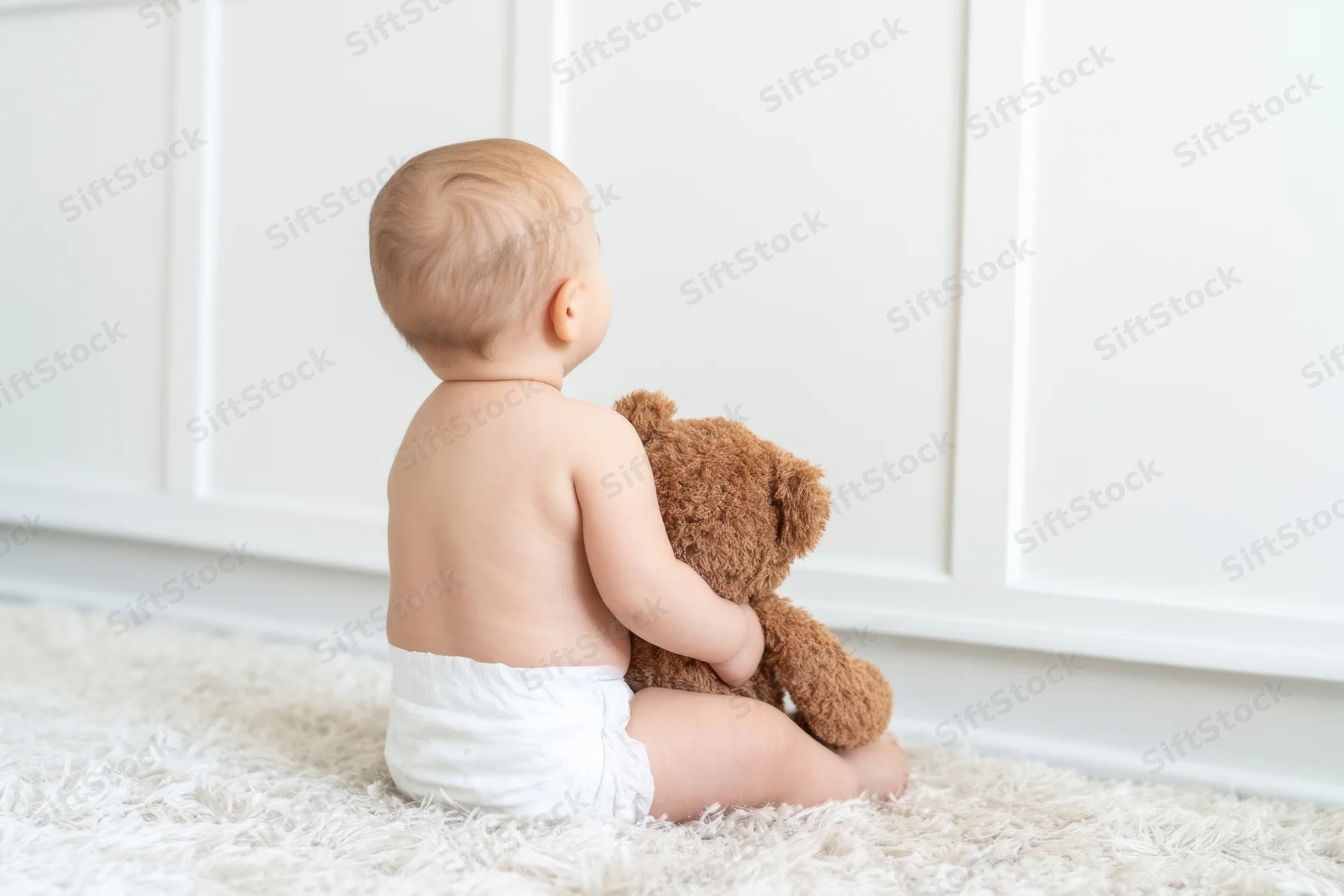 Baby sitting on light rug hugging brown teddy bear from back view