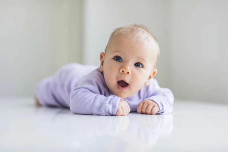 Baby lying on stomach on glossy white surface wearing purple onesie