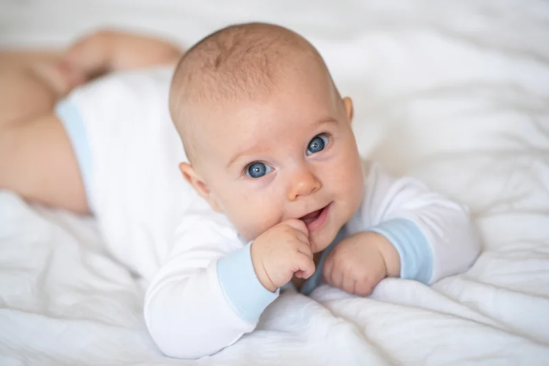 Baby lying on stomach on white blanket with hand in mouth, propped on elbows, smiling