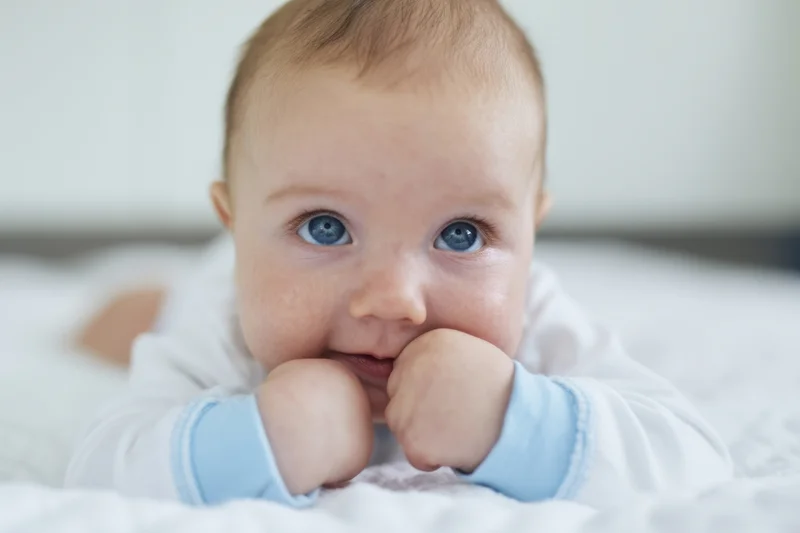 Baby lying on stomach on white blanket with hands near mouth, smiling at camera