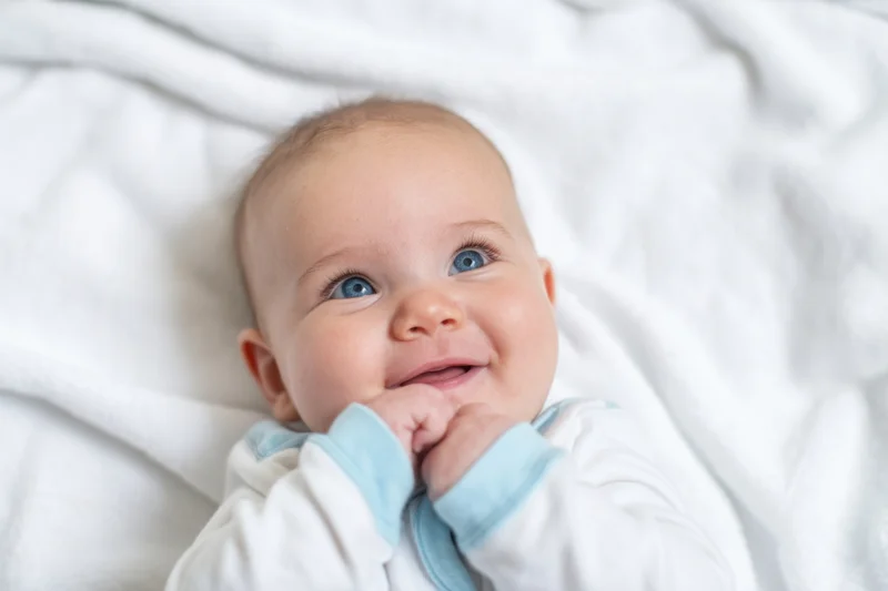 Baby lying on white blanket smiling with hands near mouth
