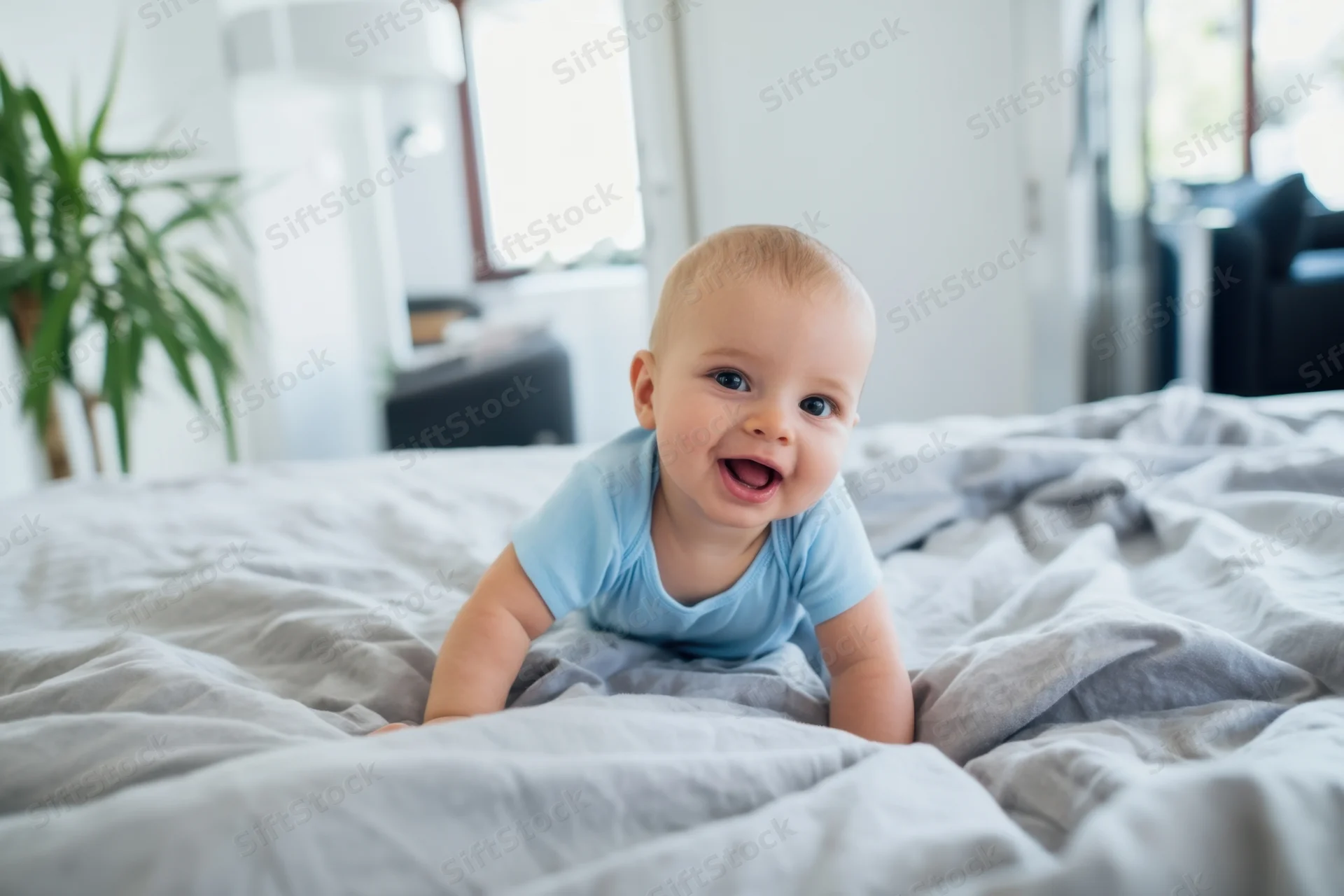 Baby crawling on soft gray comforter in bedroom, smiling with open mouth, wearing light blue onesie