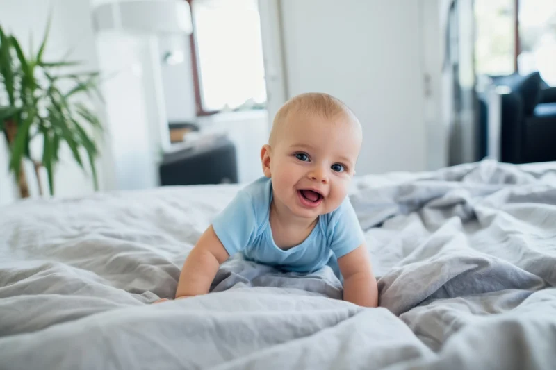 Baby crawling on soft gray comforter in bedroom, smiling with open mouth, wearing light blue onesie
