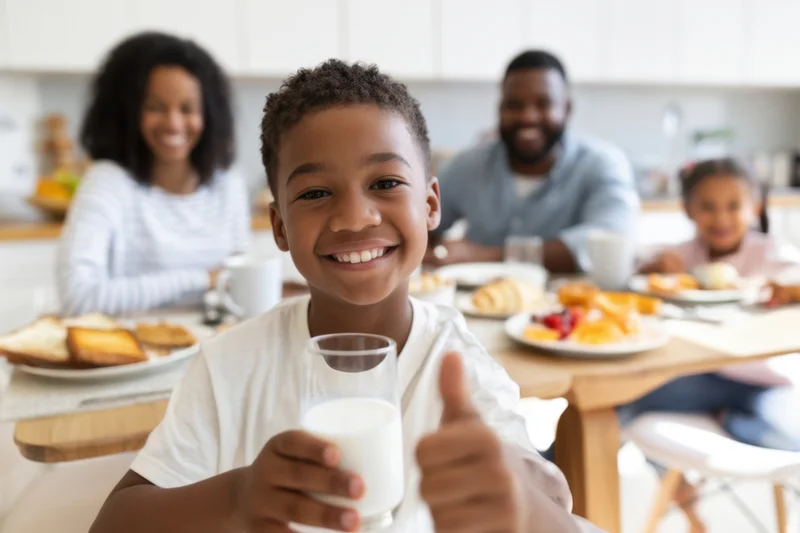 Young black boy smiling and giving thumbs up while holding a glass of milk at breakfast table with family in kitchen