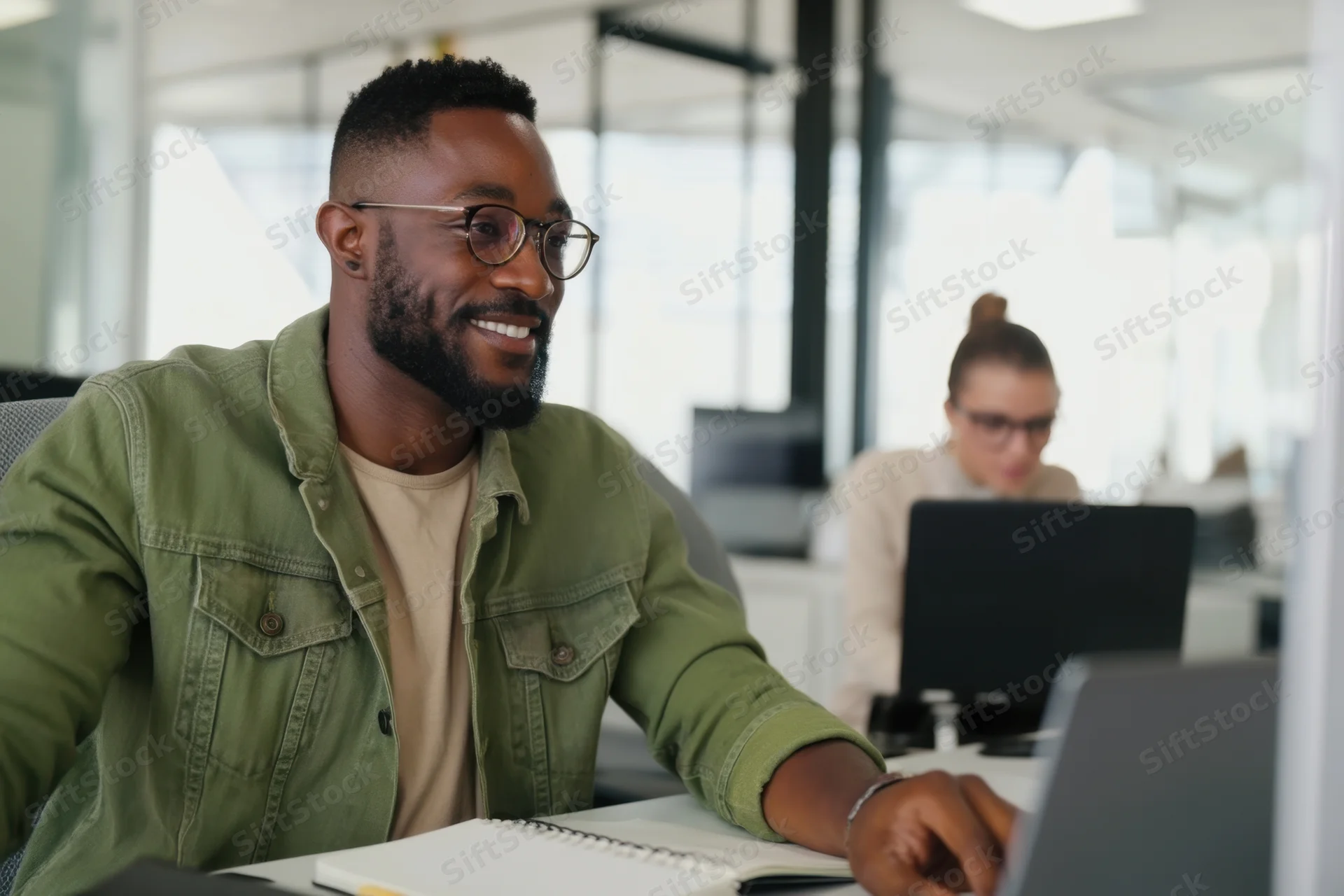 Smiling man with glasses and beard working at laptop in modern office with colleague in background