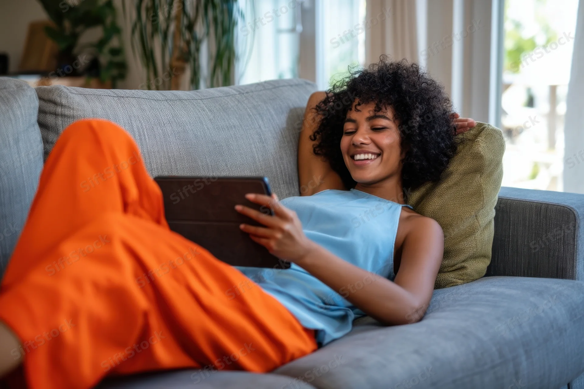 Woman smiling while lounging on gray sofa with tablet in living room