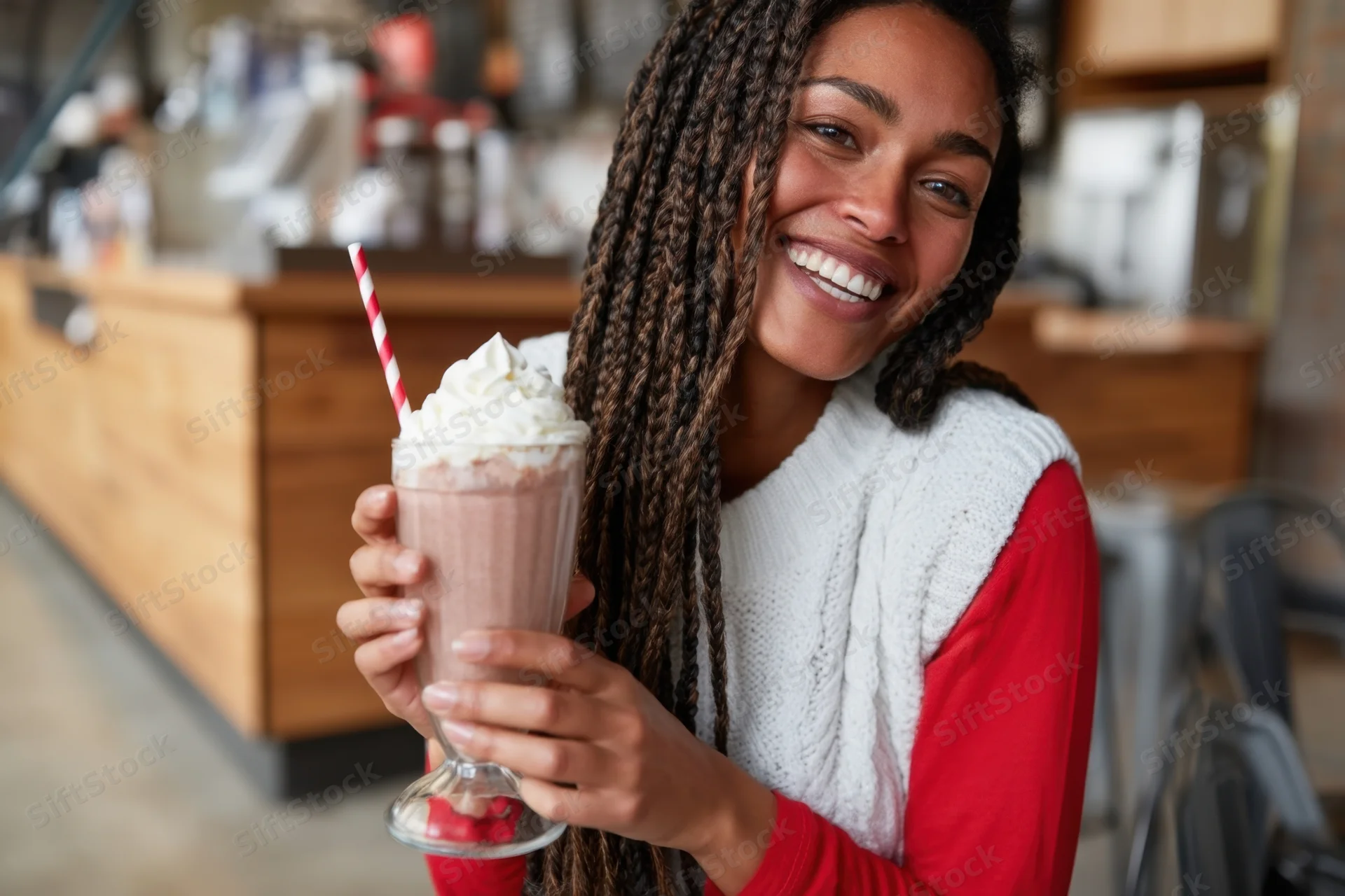 Woman smiling while holding a chocolate milkshake with whipped cream and a red-and-white straw in a cozy cafe