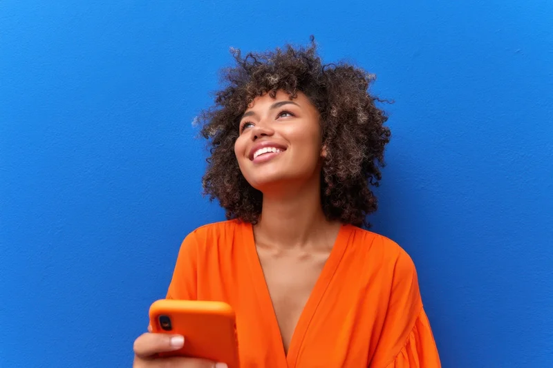 Young woman smiling while holding a phone against a blue wall