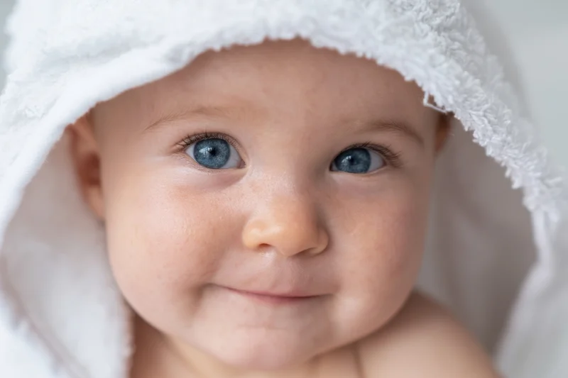 Baby with bright blue eyes peeking from a white hooded towel, smiling softly