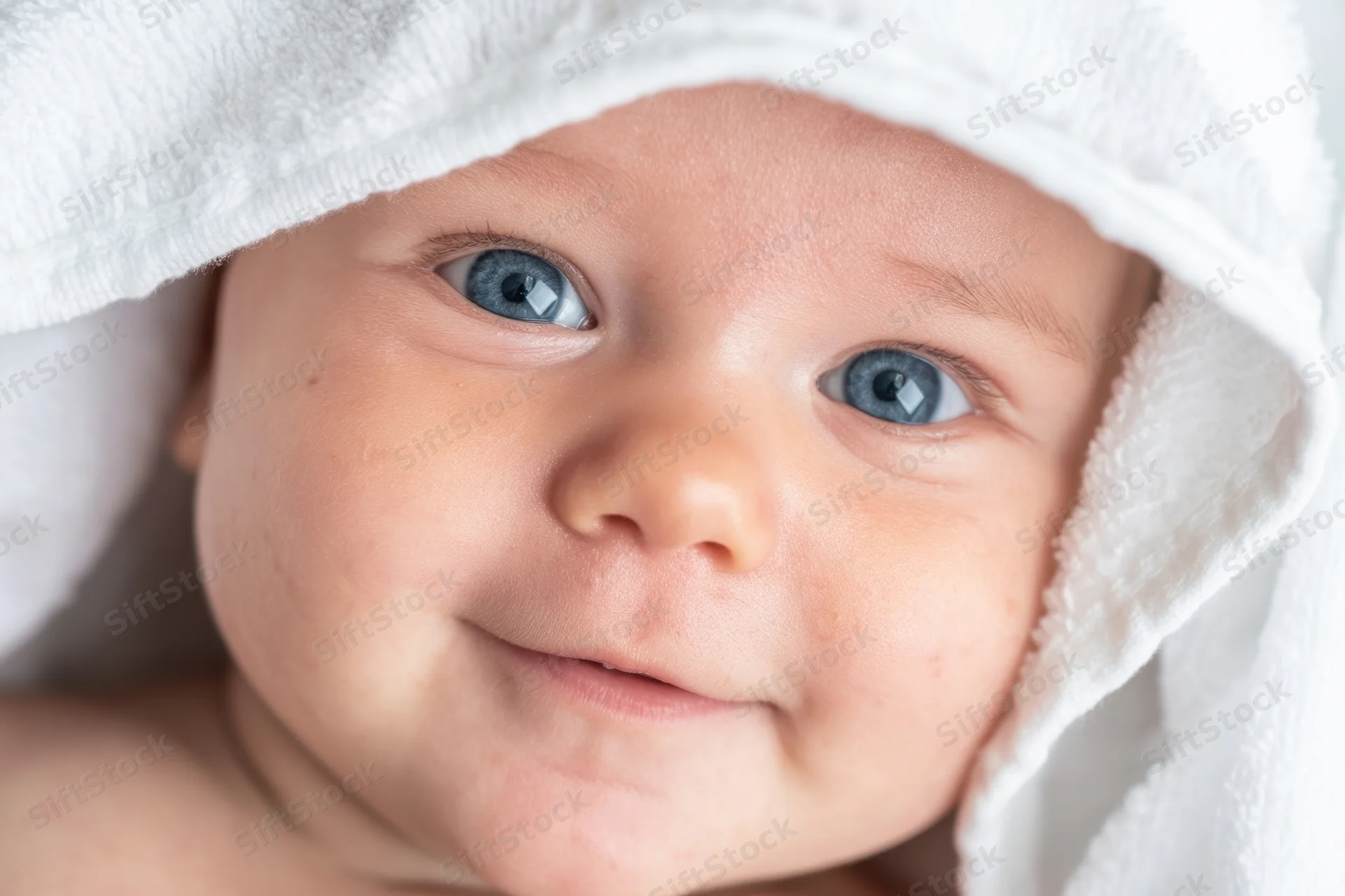 Baby with bright blue eyes peeking from a white hooded towel, smiling close-up portrait