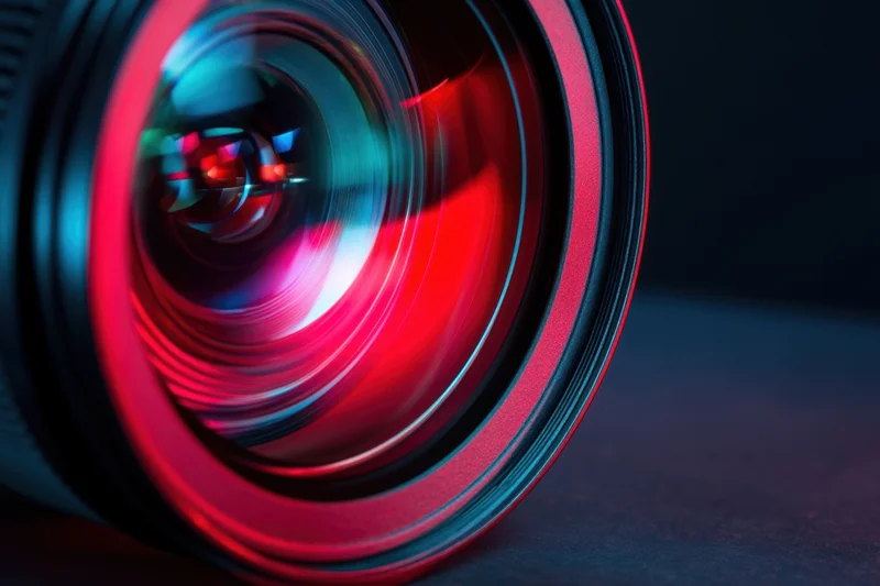 Close-up of a camera lens with neon red and blue light reflections on dark surface