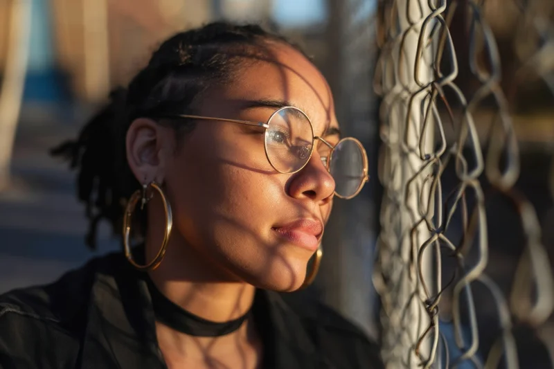 Young black woman in profile wearing round sunglasses and gold hoop earrings, leaning against chainlink fence in warm sunlight