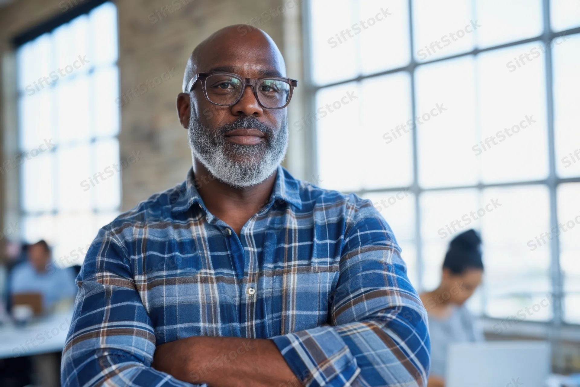 Bald man with gray beard and glasses wearing blue plaid shirt, arms crossed, in office workspace with colleagues at desks near large windows