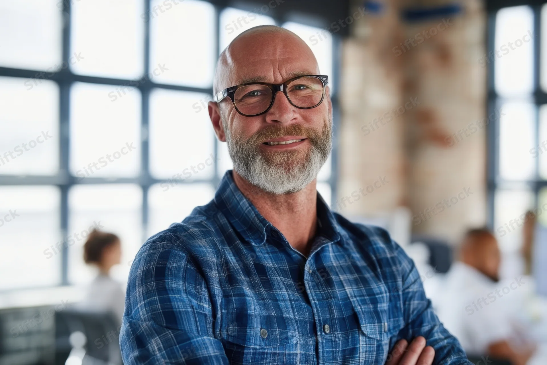 Bald man with gray beard and glasses smiling with arms crossed in modern office interior
