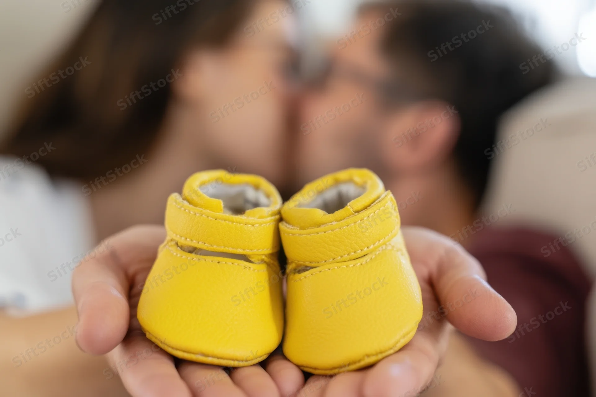 Hands holding tiny yellow baby shoes with a kissing couple in the background