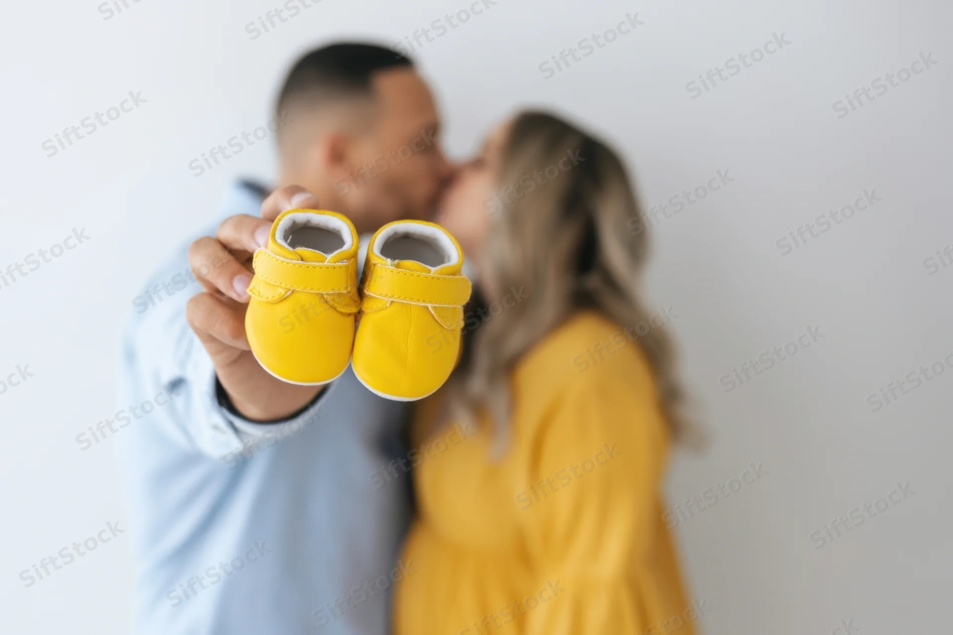 Couple kissing in the background while holding up yellow baby shoes in the foreground