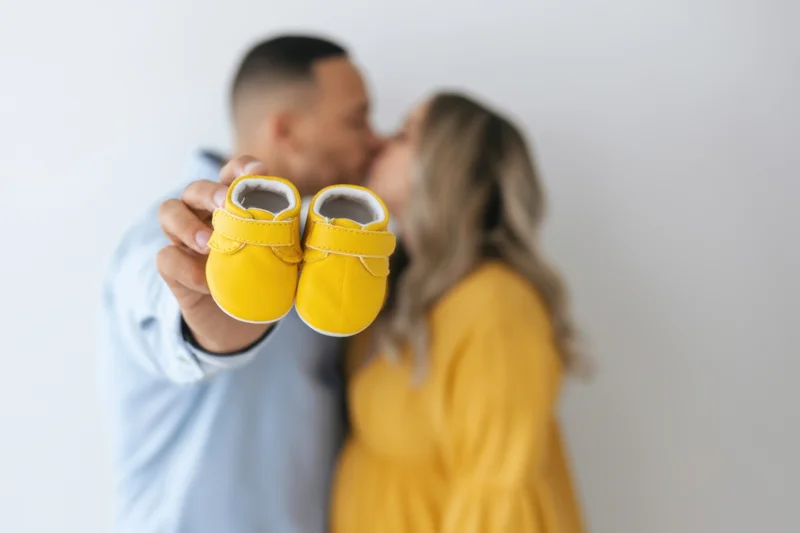 Couple kissing in the background while holding up yellow baby shoes in the foreground