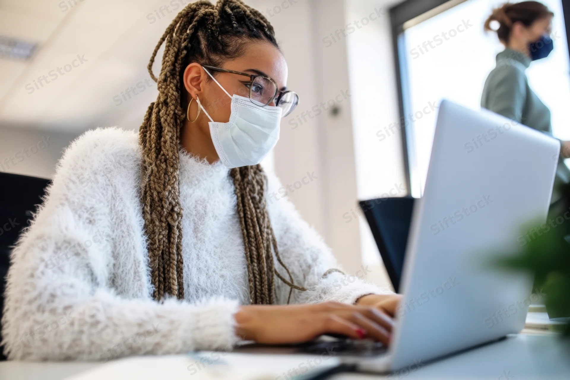 Woman with braids wearing mask and glasses typing on laptop at office desk