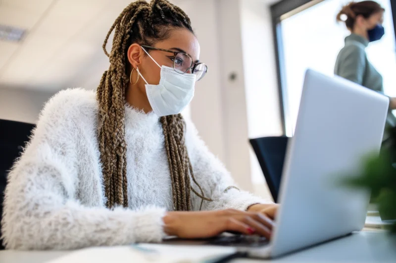 Woman with braids wearing mask and glasses typing on laptop at office desk