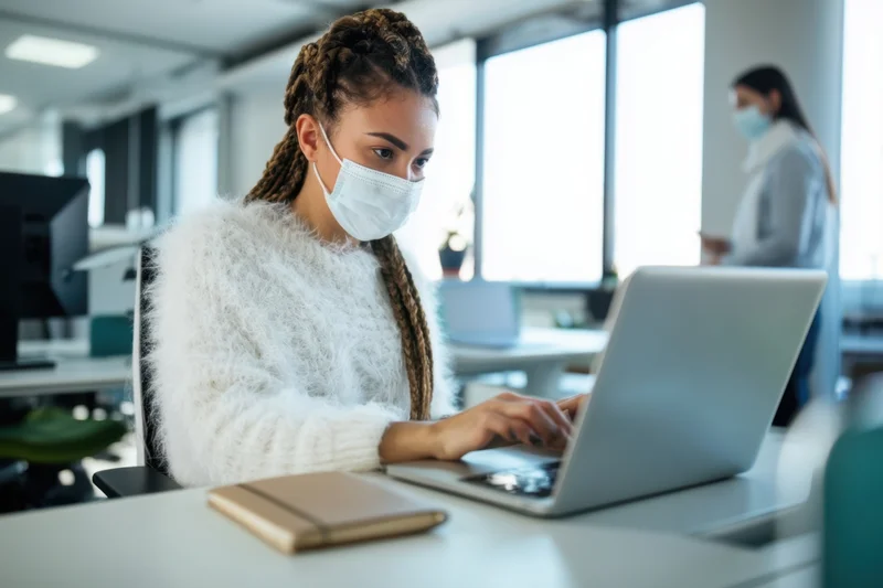 Woman typing on laptop at office desk wearing face mask
