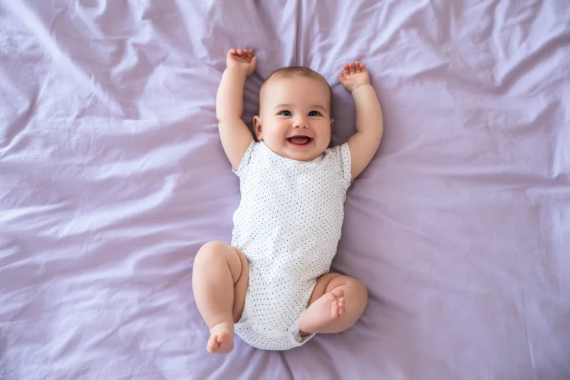 Baby lying on lavender sheet smiling with arms raised in bed