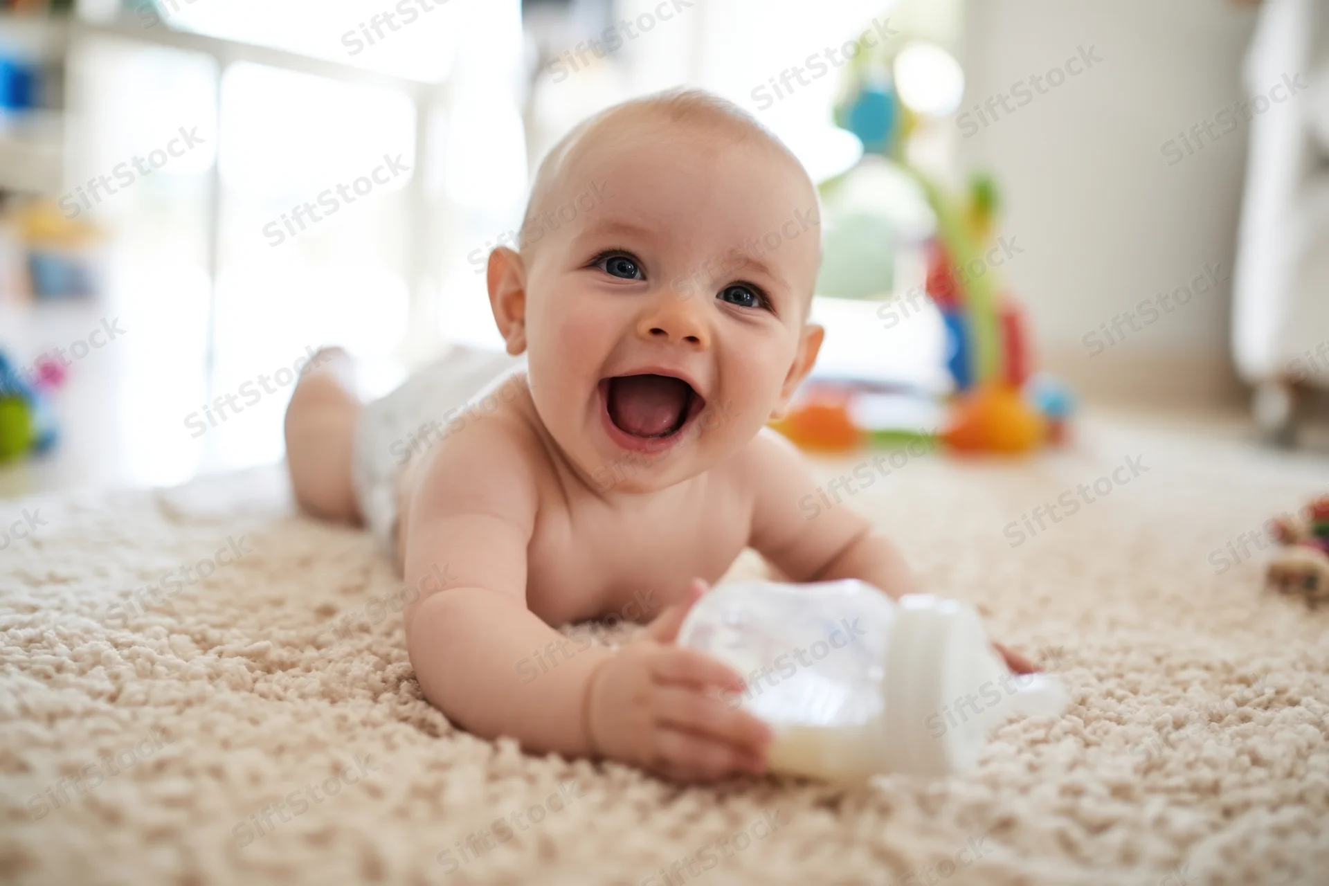 Baby lying on beige carpet smiling with bottle in hand during playtime