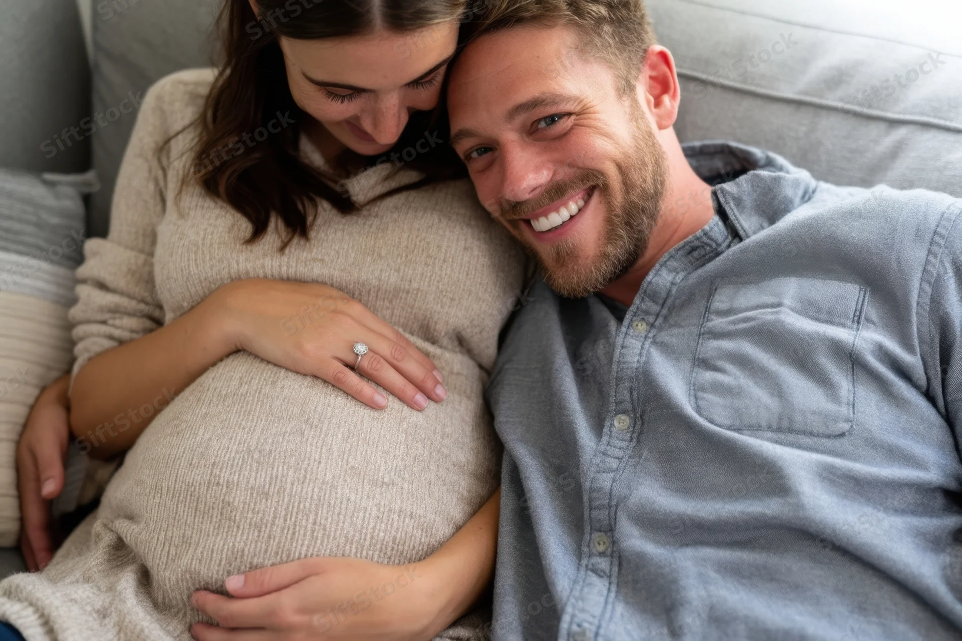 Pregnant woman resting her head on her partner’s shoulder while he smiles, both seated on a sofa at home