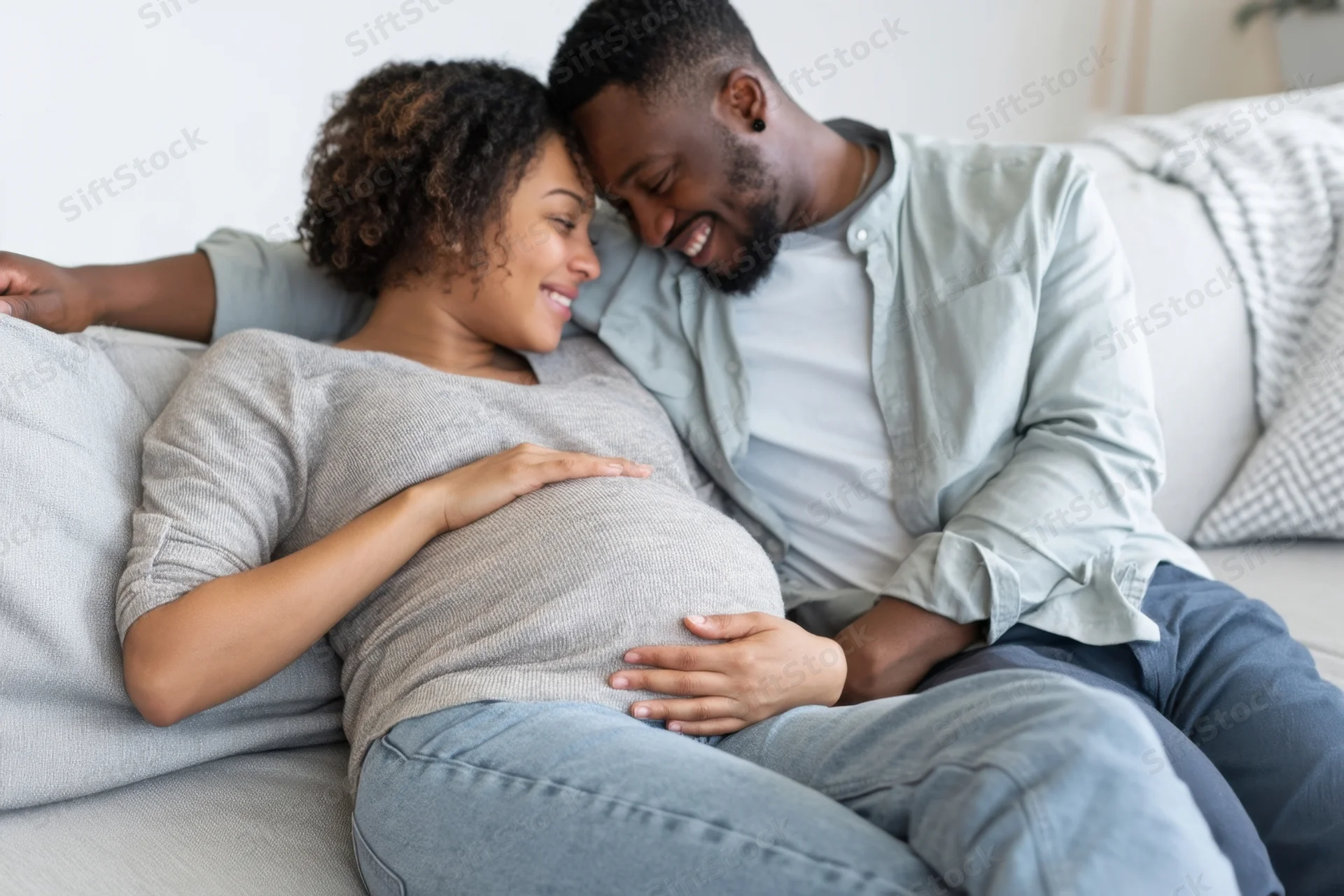 Pregnant woman and man smiling together on sofa, heads touching, hands on belly, in living room