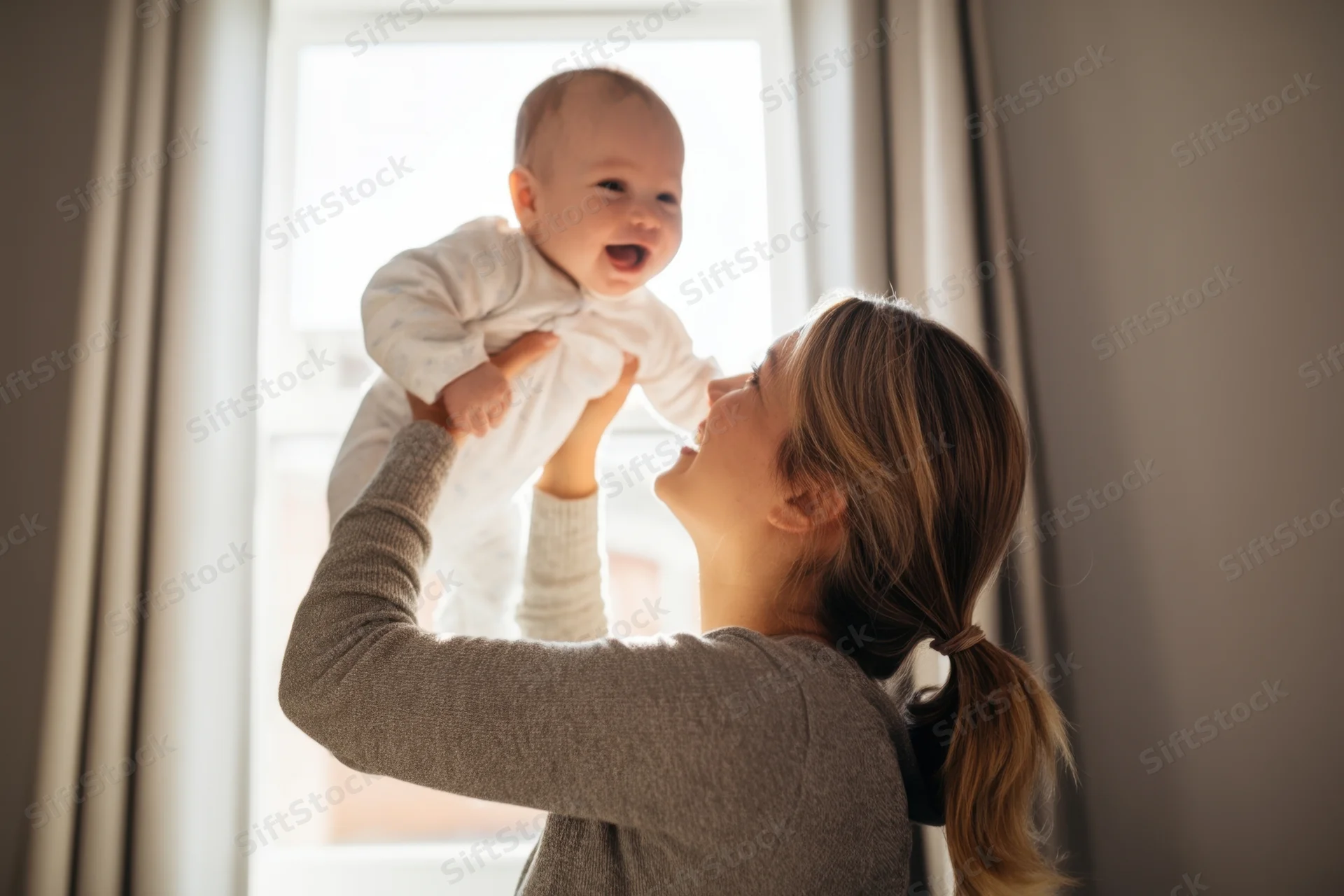 Mother lifting baby into air by window in sunlight