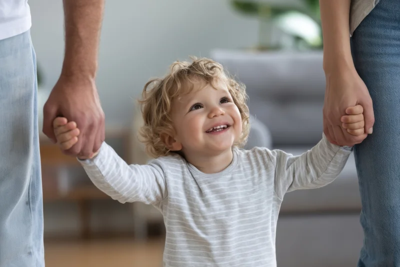 Toddler smiling while holding hands with two adults in a living room