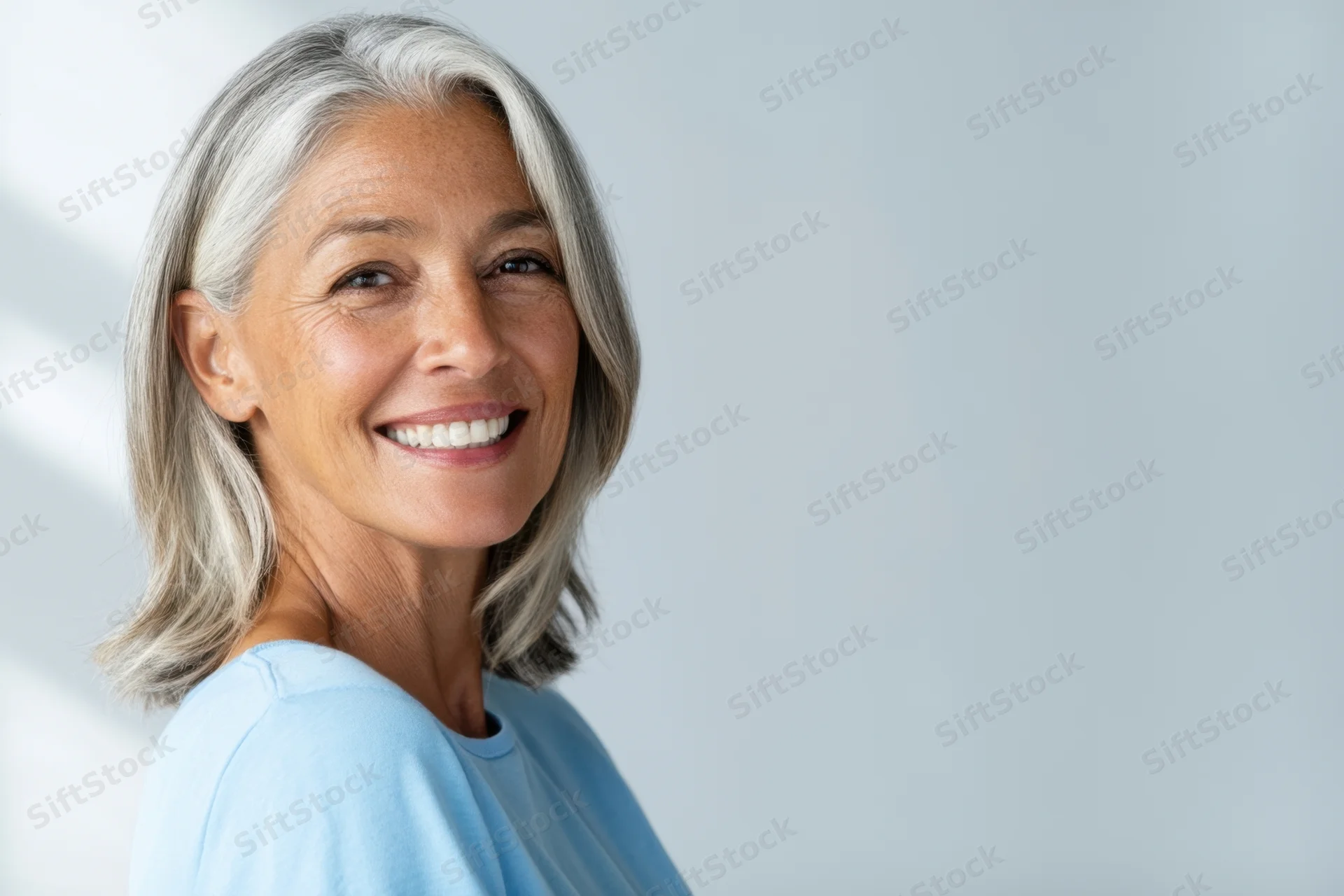 Smiling senior woman with shoulder-length gray hair in light blue shirt, side view portrait against neutral background