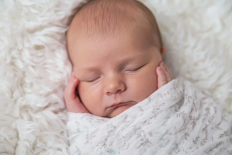 Newborn baby sleeping swaddled in a white blanket with hands near cheeks