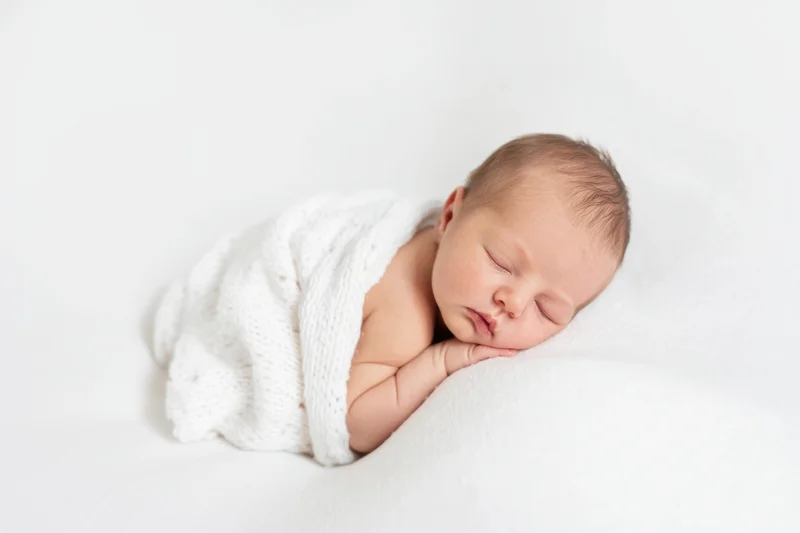 Newborn baby sleeping swaddled in white knit blanket on soft white surface