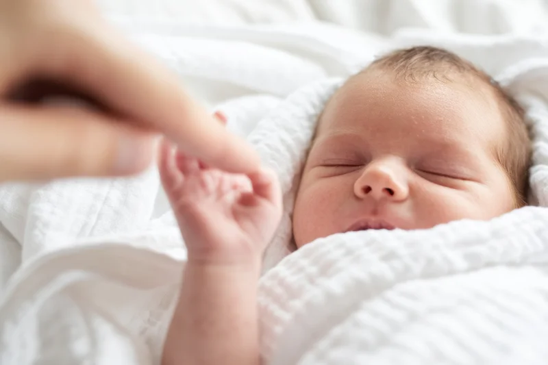 Newborn baby sleeping peacefully wrapped in white blanket with hand gently touching tiny finger