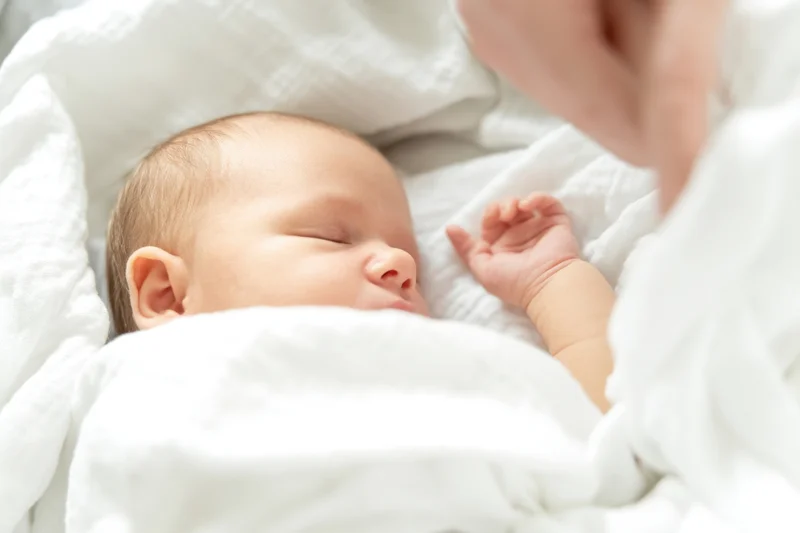 Newborn baby sleeping peacefully wrapped in white blankets with mother's hand nearby