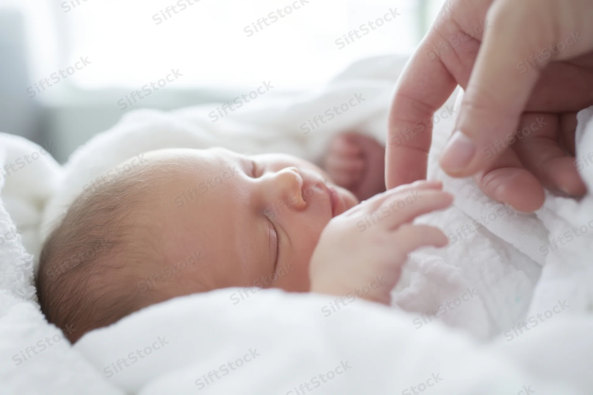 Newborn baby sleeping peacefully wrapped in white blanket with adult hand gently touching its hand