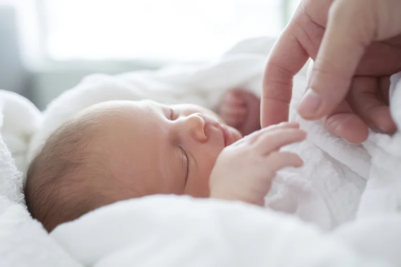 Newborn baby sleeping peacefully wrapped in white blanket with adult hand gently touching its hand
