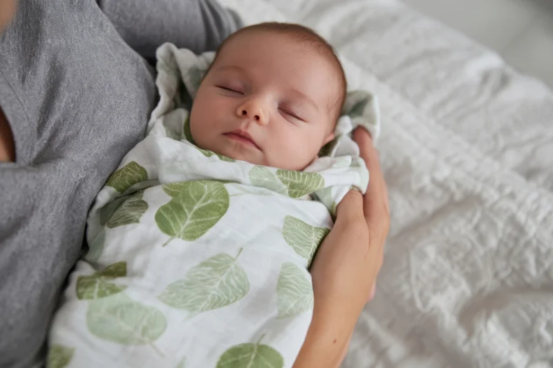Newborn sleeping swaddled in leaf-pattern blanket held by mother in bed