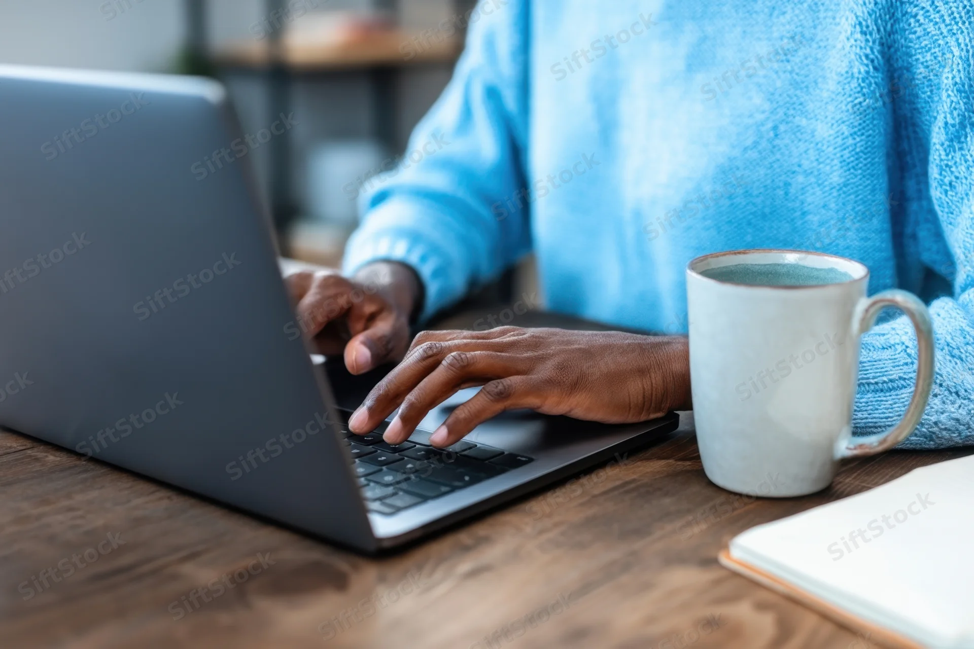 Person typing on laptop at wooden desk with mug and notebook nearby