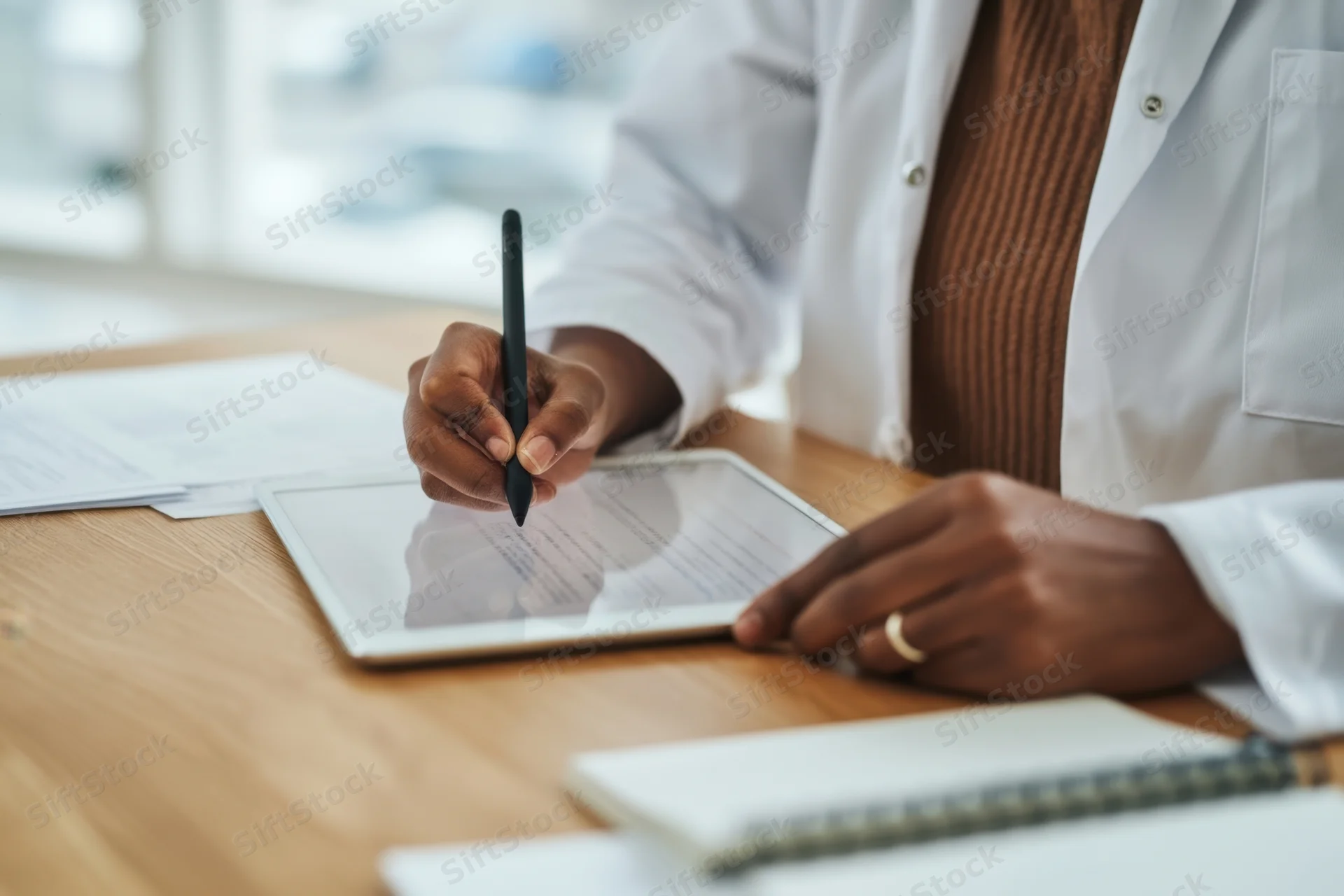Person in white lab coat writing with stylus on tablet at desk
