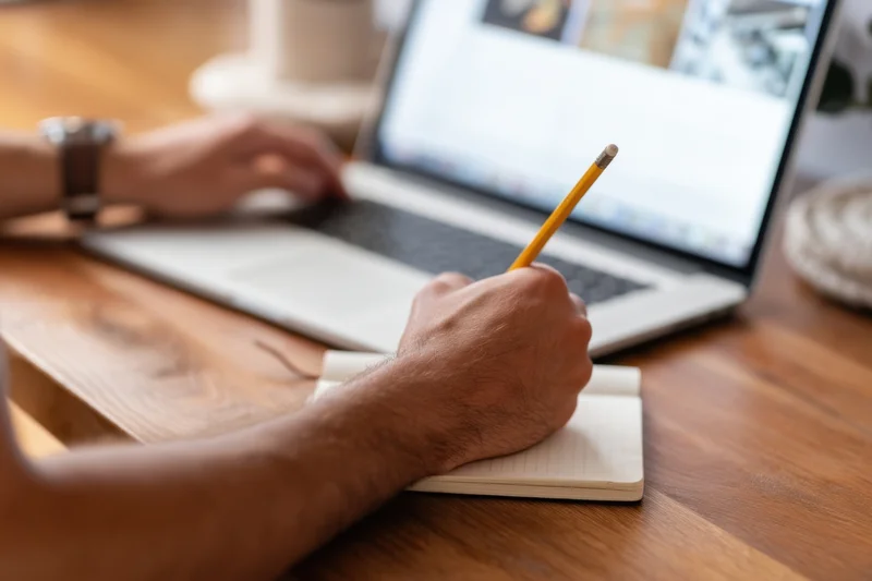 Man writing in notebook with laptop on wooden desk