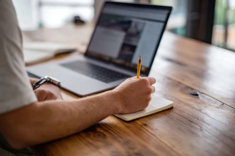 Man writing in notebook with pencil at wooden desk beside open laptop